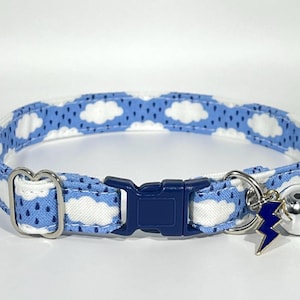 May include: A blue and white cat collar with a cloud pattern. The collar has a dark blue plastic buckle and a silver-colored metal bell with a blue lightning bolt charm. The collar is approximately 12 inches long.