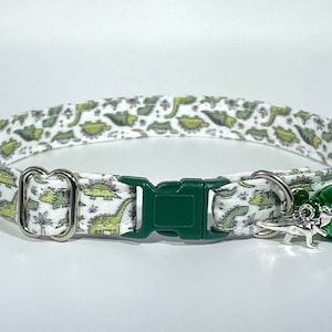 May include: A white cat collar with a green dinosaur print and a green bell. The collar has a green plastic buckle and a silver metal D-ring.