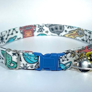 May include: A white cat collar with a colorful space-themed print, featuring rockets, planets, and stars. The collar has a blue plastic buckle and a silver bell with a planet charm.