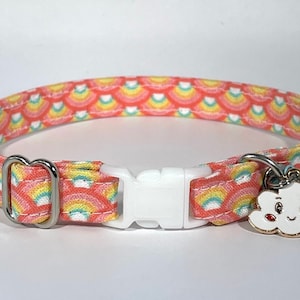 Puede incluir: Un collar para gato rosa y amarillo con un patr&oacute;n de arco&iacute;ris y una hebilla de pl&aacute;stico blanca. El collar tiene un dije adjunto en forma de una nube blanca con una cara sonriente y mejillas rojas.