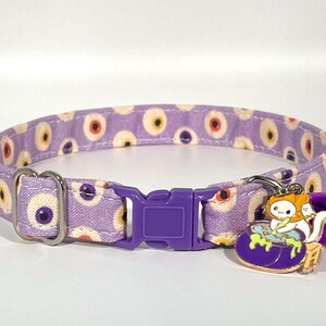 May include: Purple cat collar with a white background and a repeating pattern of colorful eyes. The collar has a purple plastic buckle and a charm of a cat stirring a cauldron with a bell.