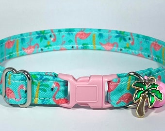 May include: A turquoise blue cat collar with a pink plastic buckle and a palm tree charm. The collar is decorated with a repeating pattern of pink flamingos and palm trees.