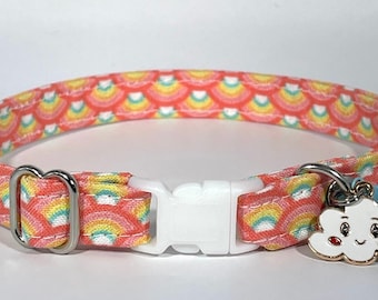 May include: A pink and yellow cat collar with a rainbow pattern and a white plastic buckle. The collar has a charm attached to it in the shape of a white cloud with a smiling face and red cheeks.