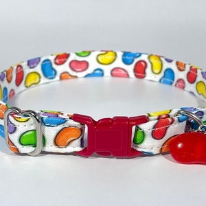 May include: A white cat collar with a colourful jelly bean pattern. The collar has a red plastic buckle and a silver bell charm with a red jelly bean charm.