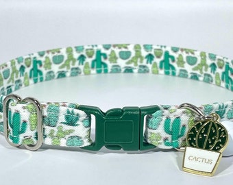 Pode incluir: Um colar de gato verde e branco com estampa de cacto e uma fivela verde. O colar tem um sino de prata e um charme que diz "Cactus".