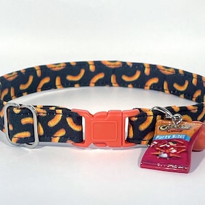 May include: A black cat collar with a repeating pattern of orange shapes. The collar has an orange plastic buckle and a silver metal loop. A small charm of a miniature bag of Cheetos is attached.