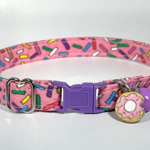 May include: Pink cat collar with a donut charm and a purple bell. The collar is covered in a sprinkle pattern.
