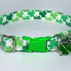 May include: A white and green cat collar with a green shamrock pattern. The collar has a green plastic buckle and a silver bell with a shamrock charm.