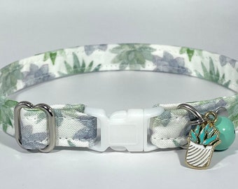 May include: A cat collar with a white plastic buckle and a silver-colored metal D-ring. The collar is patterned with green and gray succulent plants on a white background. A turquoise bell and a gold-colored charm with a succulent design are attached.