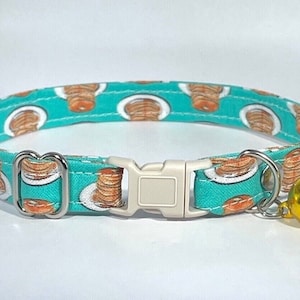 May include: A turquoise blue cat collar with a pattern of pancakes on white plates. The collar has a silver buckle and a gold bell.