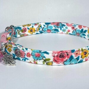 Puede incluir: Un collar de gato blanco con un patr&oacute;n floral en tonos de rosa, azul y amarillo. El collar tiene una hebilla de seguridad rosa y un charm de campana y flor plateado.