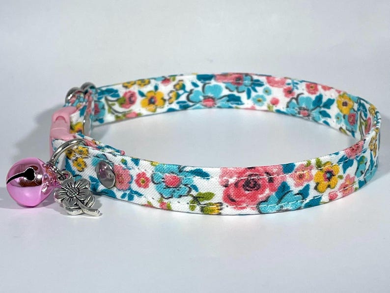 Puede incluir: Un collar de gato blanco con un patr&oacute;n floral en tonos de rosa, azul y amarillo. El collar tiene una hebilla de seguridad rosa y un charm de campana y flor plateado.