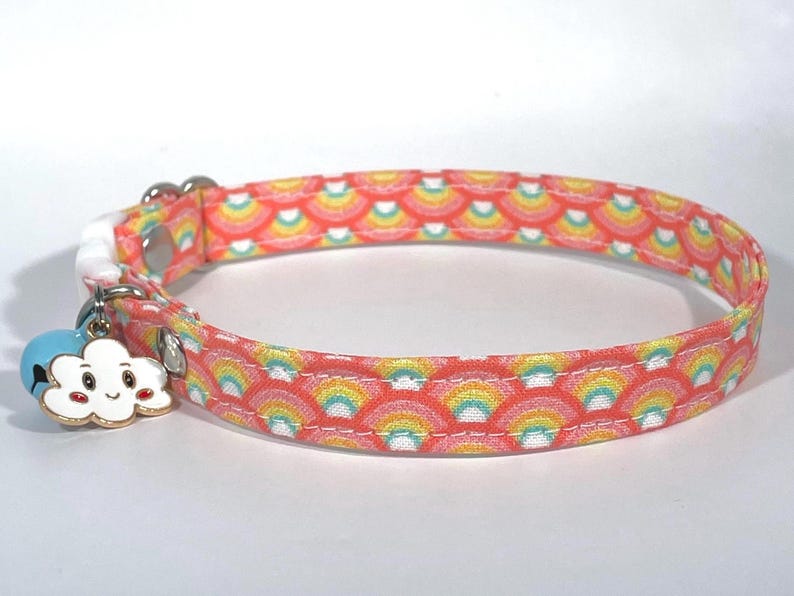 Puede incluir: Un collar para gato rosa y amarillo con un patr&oacute;n de arco&iacute;ris y un peque&ntilde;o amuleto de nube con una cara sonriente.