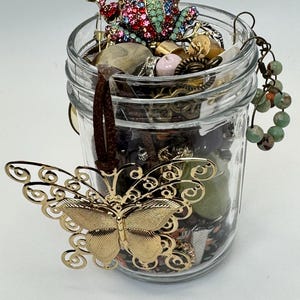 Pode incluir: Um frasco de vidro transparente cheio de joias variadas, incluindo um pingente de borboleta de metal dourado num cordão castanho, uma estatueta de sapo incrustada com strass multicoloridos e um fio de contas de pedra verde e castanha.