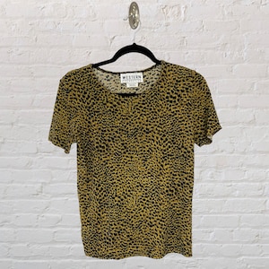 Puede incluir: Una blusa de manga corta con estampado de leopardo amarillo y negro con el texto "WESTERN CONNECTION" en la etiqueta.
