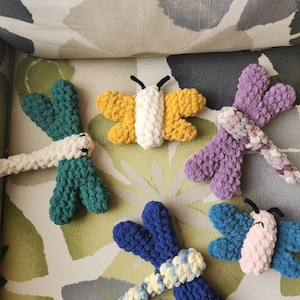 May include: Four crocheted dragonfly toys in different colors: green, yellow, purple, and blue. The toys are made with yarn and have a textured, fluffy appearance.