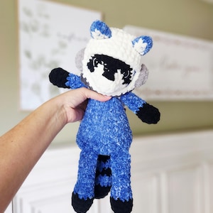 May include: A handmade crochet raccoon plush toy. The toy features a white head with blue accents, black eyes, and a black mask. The body is blue, with black paws and a striped tail. The plush is held up by a person's hand.