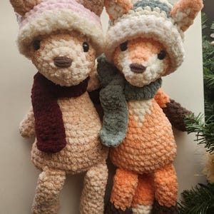 May include: Two handmade plush toys: a bear and a fox. The bear is beige with a pink and white hat and a burgundy scarf. The fox is orange and white with a blue, white, and orange hat and a green scarf. Both toys have black eyes.