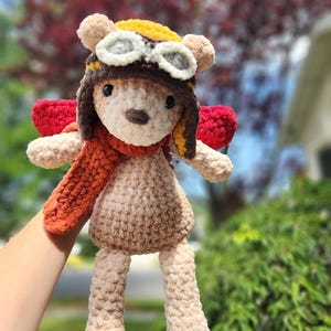May include: A handmade crochet teddy bear wearing a pilot's hat with goggles, a scarf, and red wings. The bear is light brown with a tan body and limbs. The hat is yellow and brown. The scarf is orange.