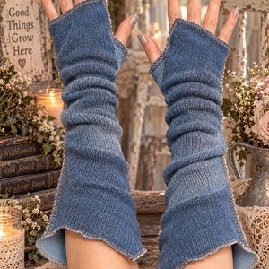 May include: A pair of blue denim fingerless arm warmers featuring a ribbed texture and exposed serged seams along the edges. These long, pull-on sleeves extend from the knuckles to the mid-forearm, showcasing a classic medium-wash blue fabric.