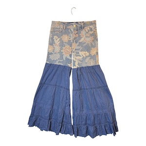 May include: High-waisted pants featuring a light blue denim upper section with a tan and white floral pattern. The lower portion consists of wide, tiered blue fabric legs with horizontal lace trim detailing and a ruffled hemline at the bottom.