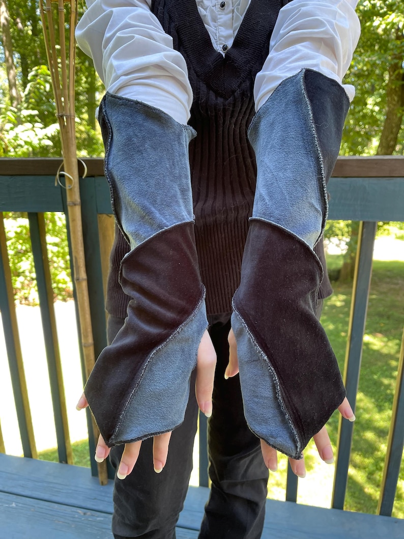 May include: A pair of fingerless arm warmers featuring a color-blocked design of dark charcoal gray and light blue fabric panels. The sleeves have exposed seams and a diagonal construction that extends from the wrist up to the mid-forearm area.