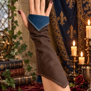 May include: A long, fingerless glove in dark brown fabric with a contrasting blue inner lining. The glove features exposed orange stitching along the seams and a pointed cuff design that extends over the back of the hand and down the forearm.