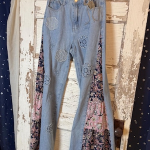 May include: Light blue denim jeans featuring a high-waisted fit, a button closure, and frayed flower-shaped patches on the front. The sides are accented with tiered, multi-colored floral fabric panels in shades of pink, navy, and white.