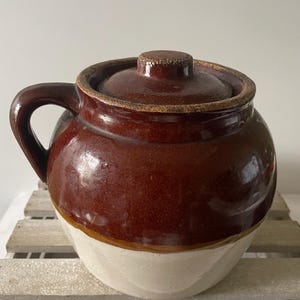 May include: A brown and cream-colored ceramic crock with a lid and handle. The crock has a rounded shape with a dark brown top and lid, and a cream-colored bottom. The handle is arched and attached to the side.