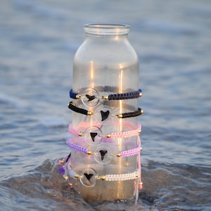 May include: A clear glass bottle with a stack of colorful bracelets featuring shark tooth charms. The bracelets are made of braided cord and have gold beads. The bottle is sitting in the water.