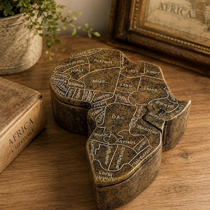 Peut inclure: Une boîte décorative en forme de continent africain avec une finition bronze texturée et mouchetée. Le couvercle présente une carte gravée de l'Afrique avec les noms des pays inscrits en blanc, notamment l'Algérie, le Niger, la Libye et l'Égypte.