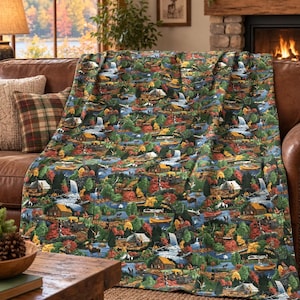 May include: A throw blanket featuring a detailed outdoor scene with cabins, tents, canoes, waterfalls, and wildlife set against a backdrop of dense green and autumn-coloured trees. The pattern includes various figures engaged in camping activities.