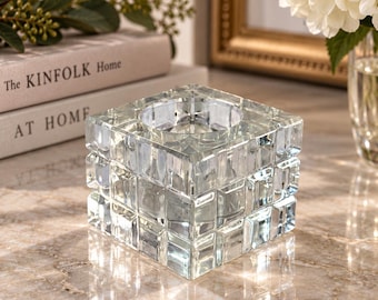 Puede incluir: Un portavelas cuadrado de cristal construido a partir de una rejilla de pequeños bloques de cristal transparentes en forma de cubo. El objeto presenta un centro circular hueco diseñado para sostener una vela, con una superficie facetada.