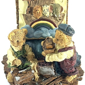 May include: A detailed figurine depicting Noah's Ark with two teddy bears. The ark features a giraffe, lion, zebra, and elephant. A rainbow arches over the ark. The bears are dressed in work clothes, with one holding a tool. The base is textured.