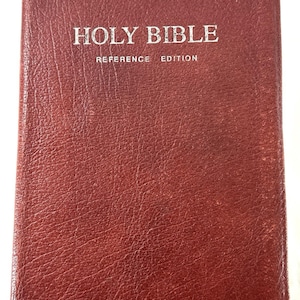 May include: A closed, red leather-bound Holy Bible with the words "HOLY BIBLE REFERENCE EDITION" in silver lettering. The name "Karl D. Britt" is printed in silver at the bottom of the cover. The book has a textured surface.