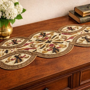 May include: An ornate, beige and brown table runner with a repeating floral pattern. The runner is placed on a wooden surface, with a vase of white flowers and two antique books in the background.