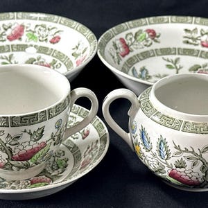 Może przedstawiać: Biały ceramiczny zestaw do herbaty z kwiatowymi i geometrycznymi wzorami. Zestaw zawiera dwie miski, filiżankę ze spodkiem i cukiernicę. Kwiatowy wzór ma różowe, zielone i niebieskie akcenty.