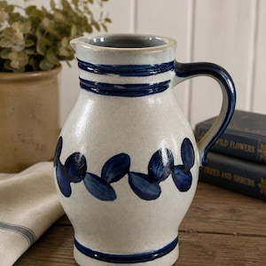 May include: A ceramic pitcher with a cream-colored base and blue accents. The pitcher features a dark blue handle and is decorated with blue leaf-like patterns. Three horizontal blue stripes encircle the top.