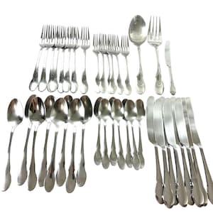 Puede incluir: Una gran colección de cubiertos de acero inoxidable que incluye varios tenedores, cucharas y cuchillos. El conjunto presenta utensilios de metal plateado pulido con mangos simples y lisos, dispuestos en filas organizadas sobre un fondo blanco.