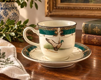May include: A cream-coloured ceramic teacup and saucer set featuring a mallard duck illustration. The rim is decorated with a dark teal band, gold filigree accents, and delicate gold dotted lines. The handle is curved and white.