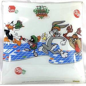 May include: Square glass dish featuring Looney Tunes characters. Bugs Bunny, Daffy Duck, Marvin the Martian, and the Tasmanian Devil are depicted in a colorful design. The dish has a blue and white checkered pattern and the words "Trick Chase".