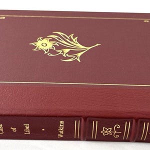 Pode incluir: Um livro de cor bordô com desenhos florais e geométricos dourados. A capa apresenta um emblema floral dourado e uma borda dourada. A lombada mostra o título "A Slight Case of Libel" e o nome do autor, Watkins, em letras douradas. Uma fita vermelha de marcador é visível.