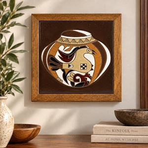 May include: A square ceramic tile framed in wood, featuring a dark brown background with a central graphic of a stylized bird inside a vessel shape. The design uses cream, orange, and black colors with geometric patterns on the vessel rim.