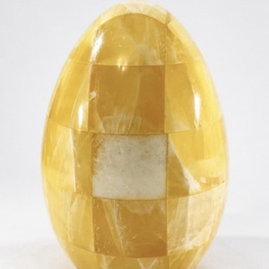 May include: A decorative egg-shaped object with a mosaic pattern. The egg is composed of yellow and off-white rectangular pieces, creating a checkerboard effect. The surface has a glossy finish, reflecting light.