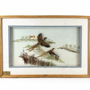 Könnte beinhalten: Gerahmtes Kunstwerk mit zwei fliegenden Fasane über einer Landschaft mit einem kleinen Haus. Das Kunstwerk ist in einem goldfarbenen Holzrahmen mit hellblauem Rand gefasst. Die Fasane sind braun und hellbraun.