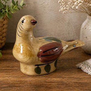 May include: A ceramic bird ornament in tan, brown, and green. The bird features a detailed design with a red beak and wing and tail accents. The ornament is displayed on a wooden surface.