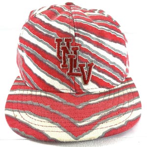 Puede incluir: Una gorra de béisbol roja, blanca y gris con un patrón de rayas. La gorra presenta las letras "UNLV" en bordado rojo en la parte delantera. La visera también es roja, blanca y gris.