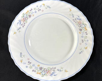 May include: A white ceramic dinner plate with a scalloped edge and a light blue rim. The plate features a delicate floral pattern with small pink, yellow, purple, and blue flowers along with green leaves arranged in clusters around the border.