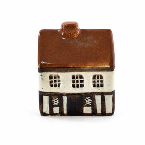 Puede incluir: Una figura de casa de cerámica con techo y chimenea marrones, una sección media blanca con tres ventanas cuadradas y una base a rayas blancas y negras. La casa en miniatura es un objeto decorativo.