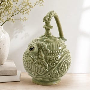 May include: A sage green ceramic vessel with a rounded body and a high arched handle. The surface features intricate relief carvings of floral motifs and a sculpted lion-like figure near the spout. The glaze has a glossy, smooth finish.